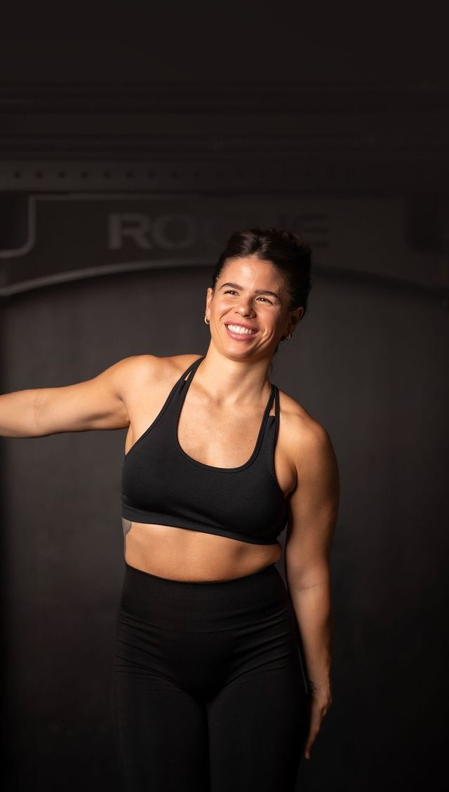 A woman in a black sports bra and black pants is standing in front of a black wall.