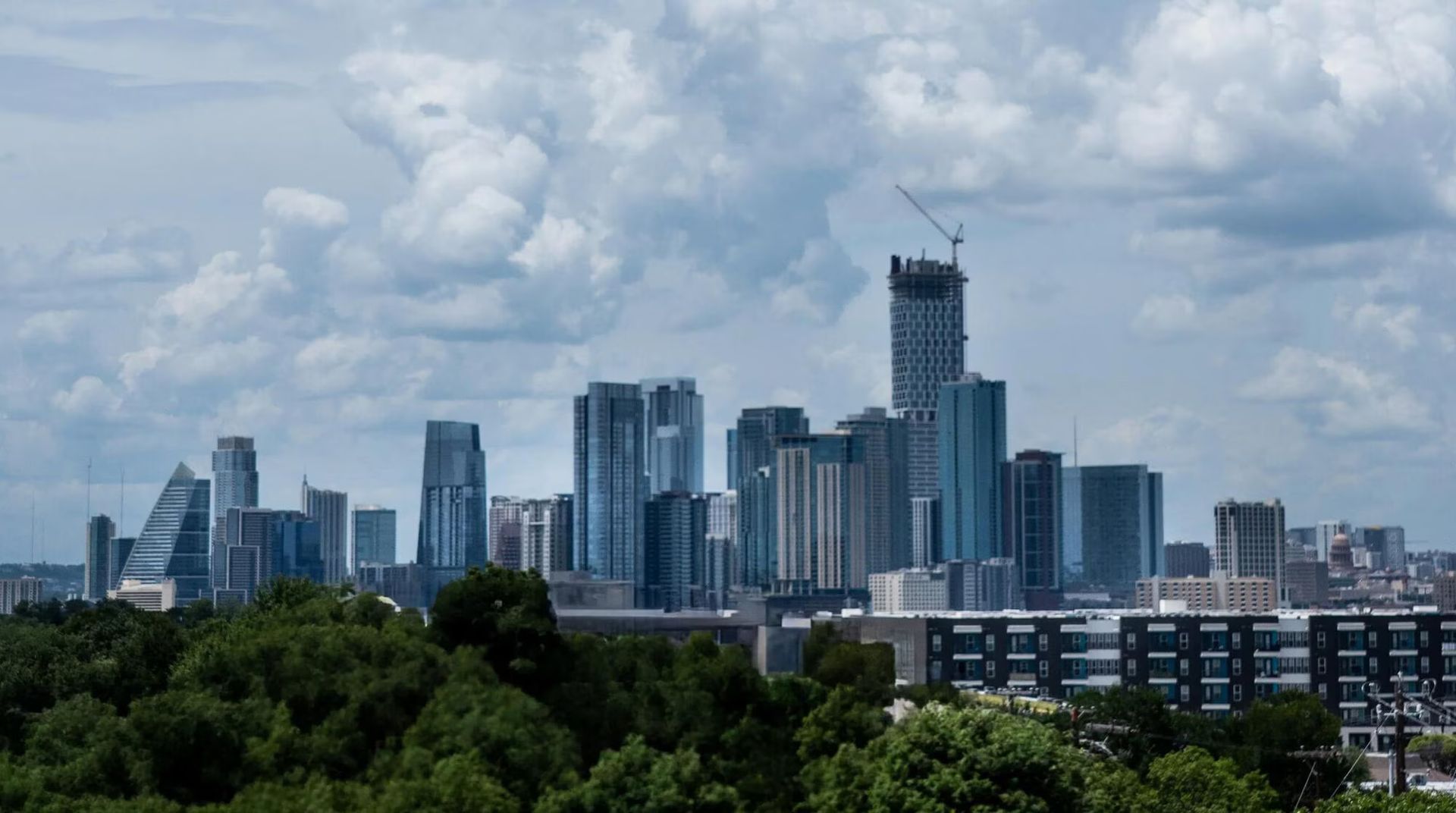 City skyline with tall buildings under a cloudy sky, trees in the foreground.