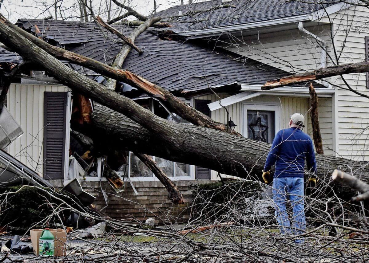 A man is standing in front of a house that has been damaged by a tree.