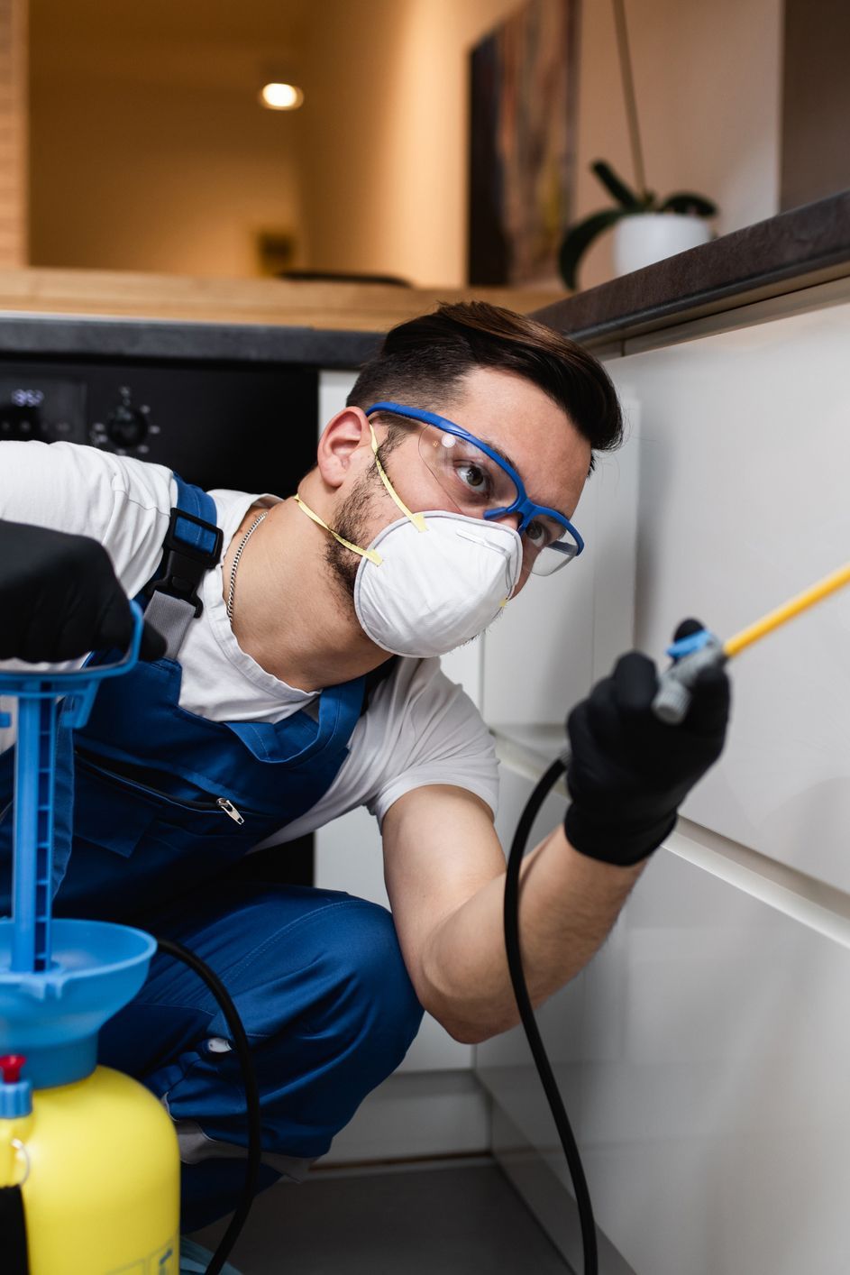 A man wearing a mask and safety glasses is spraying a chemical on a cabinet in a kitchen.