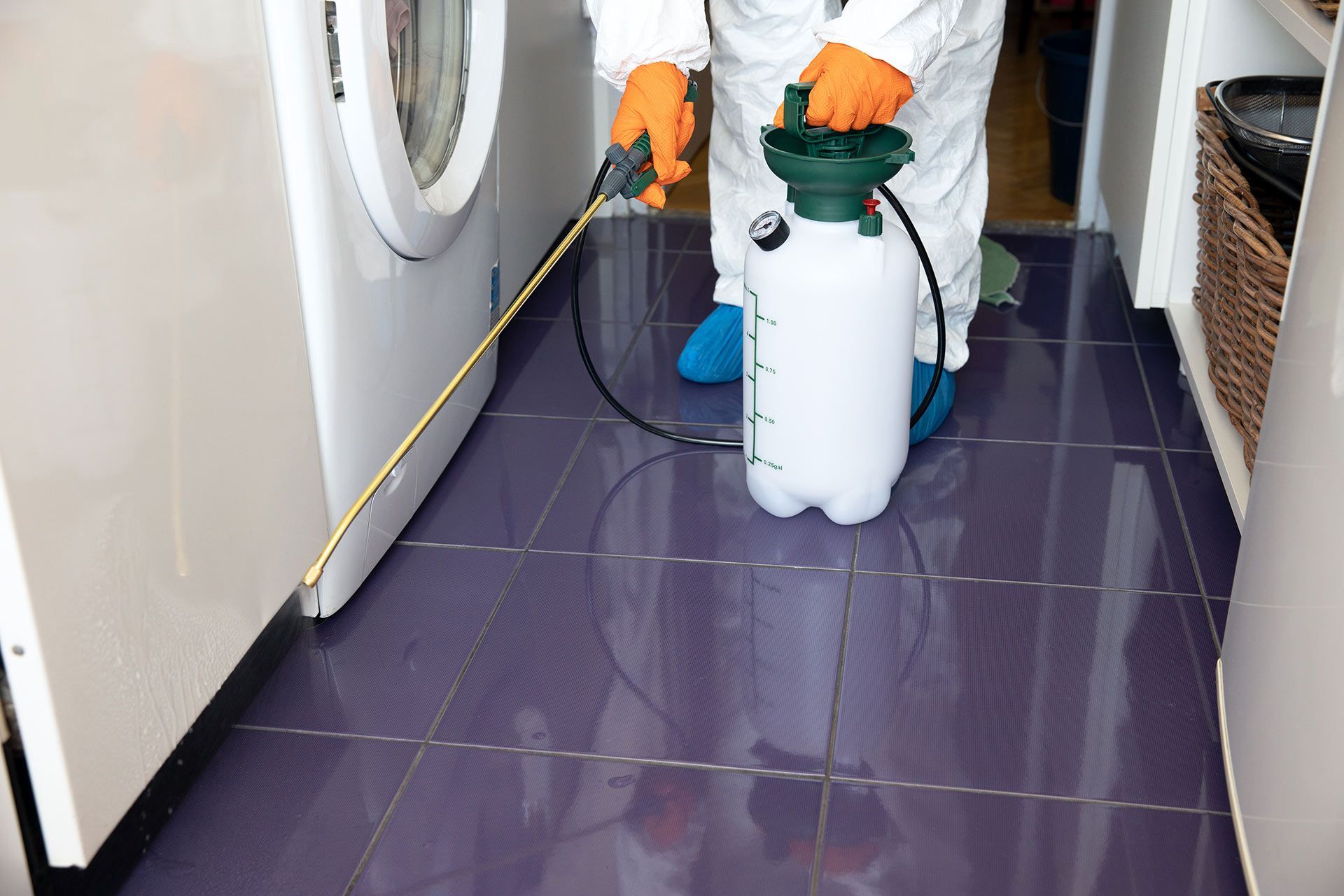 A pest control worker sprays treatment along the tiled floor near the washing machine.