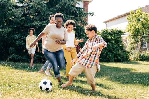 Family playing soccer in a grassy yard. Woman and boy kick the ball; others watch. Sunny day.