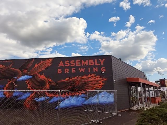 Assembly Brewing building with orange and black mural of a bird, gray exterior, cloudy sky.