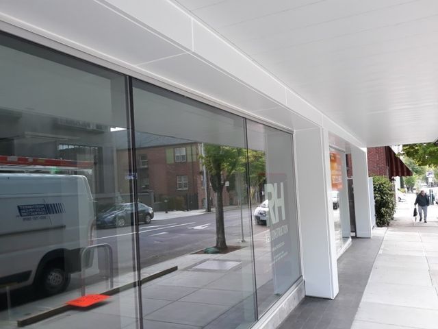 Exterior of a white storefront with large glass windows reflecting the street. A person walks on the sidewalk.