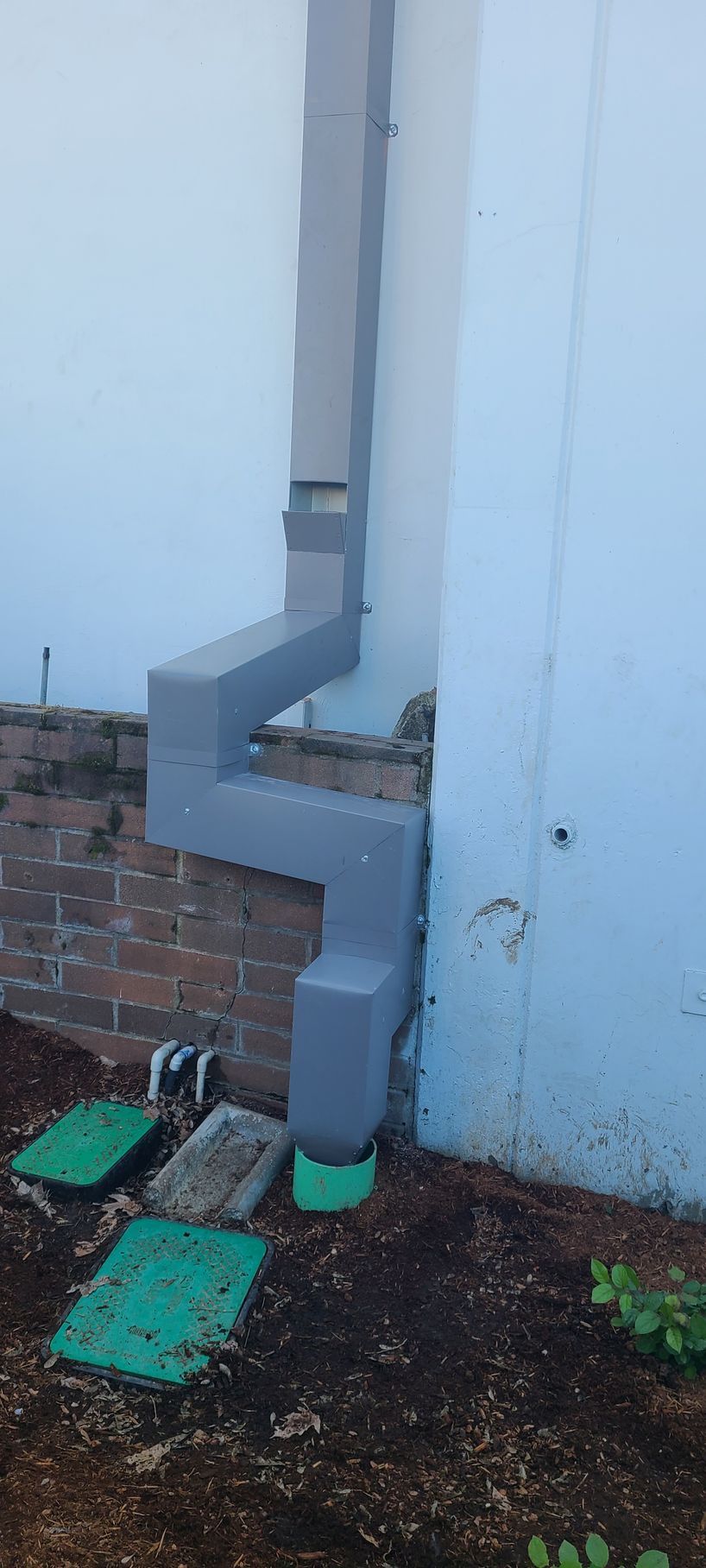Downspout with a cleanout installed near the ground, against a light blue wall and brick foundation.