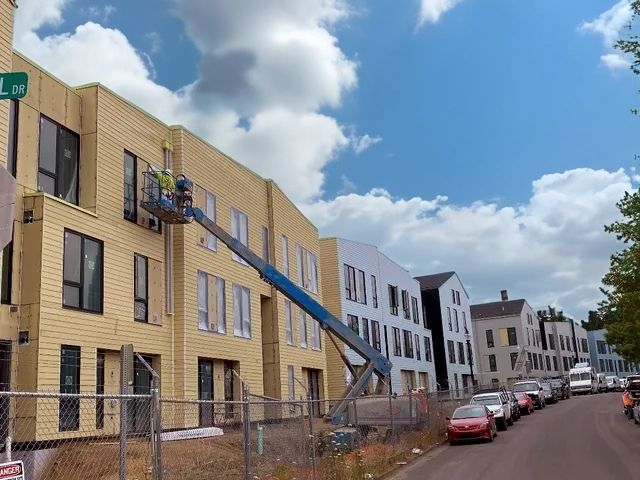 Gresham Station Apartments under construction, workers in lift, yellow and blue buildings, cars parked on street.