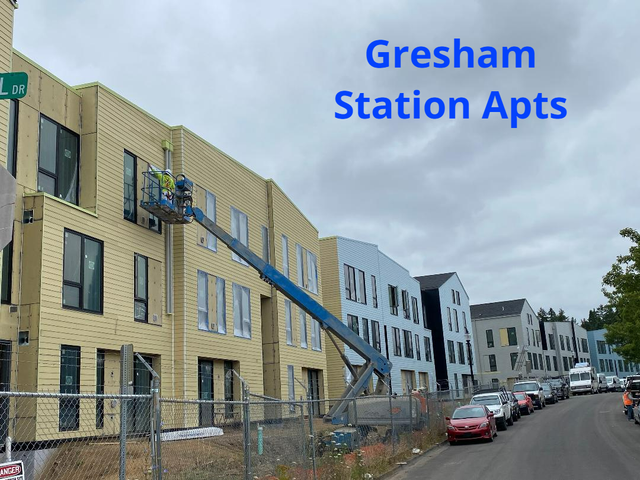 Gresham Station Apartments under construction, workers in lift, yellow and blue buildings, cars parked on street.
