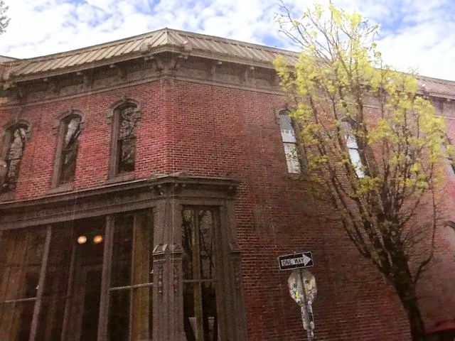 Brick building with ornate woodwork, corner view. Tree on the right, street sign in front. Overcast day.