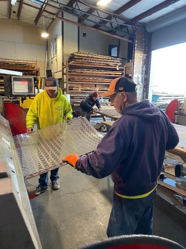 Three workers handling a large metal sheet in a workshop. One in yellow, one in purple, and one in the background.
