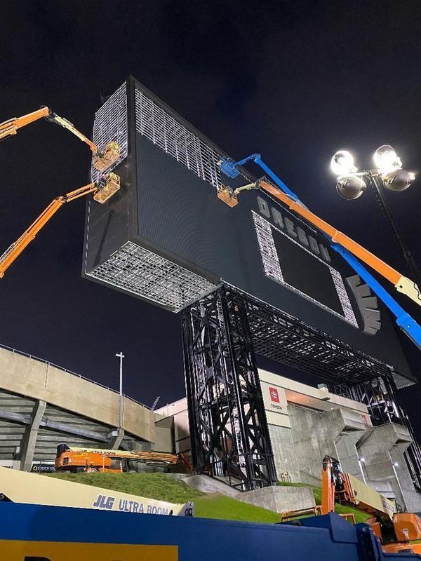 Large stadium scoreboard being installed at night with assistance of aerial lifts.