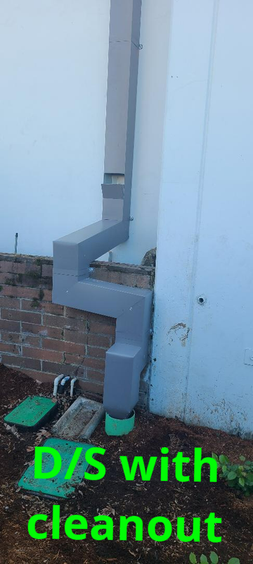 Downspout with a cleanout installed near the ground, against a light blue wall and brick foundation.