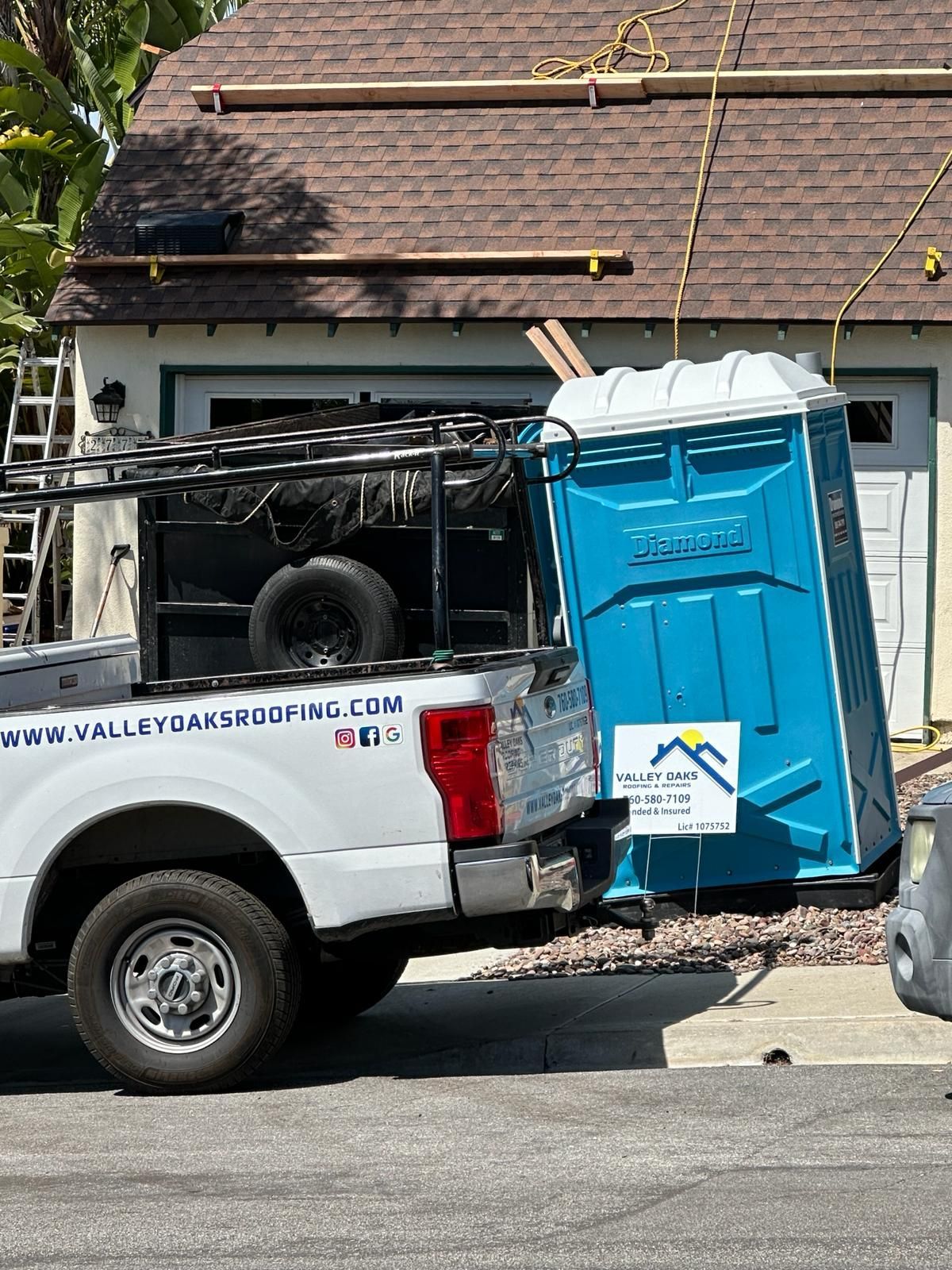 A white truck with a blue portable toilet in the back is parked in front of a house.