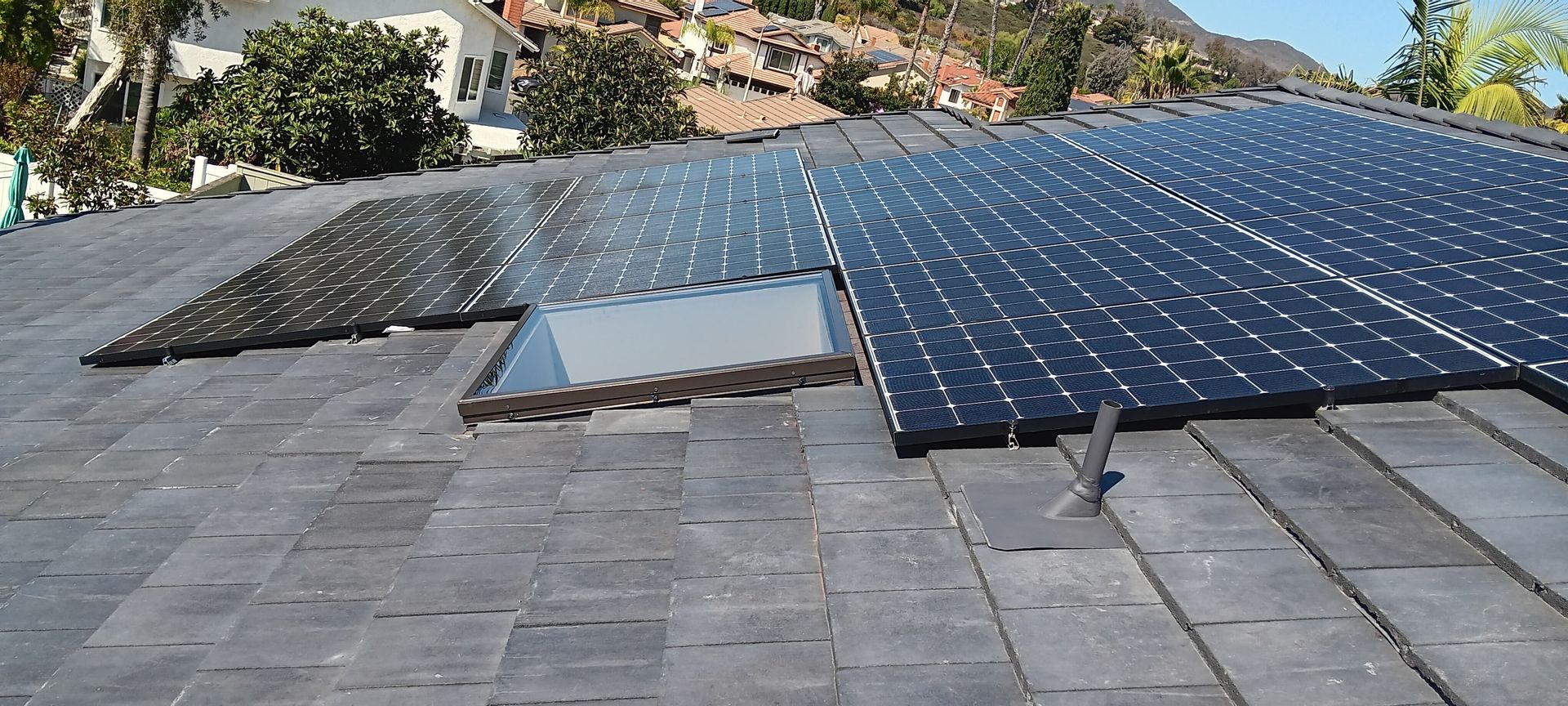 Solar panels on a slate roof with a skylight, sunny day, trees and houses in the background.