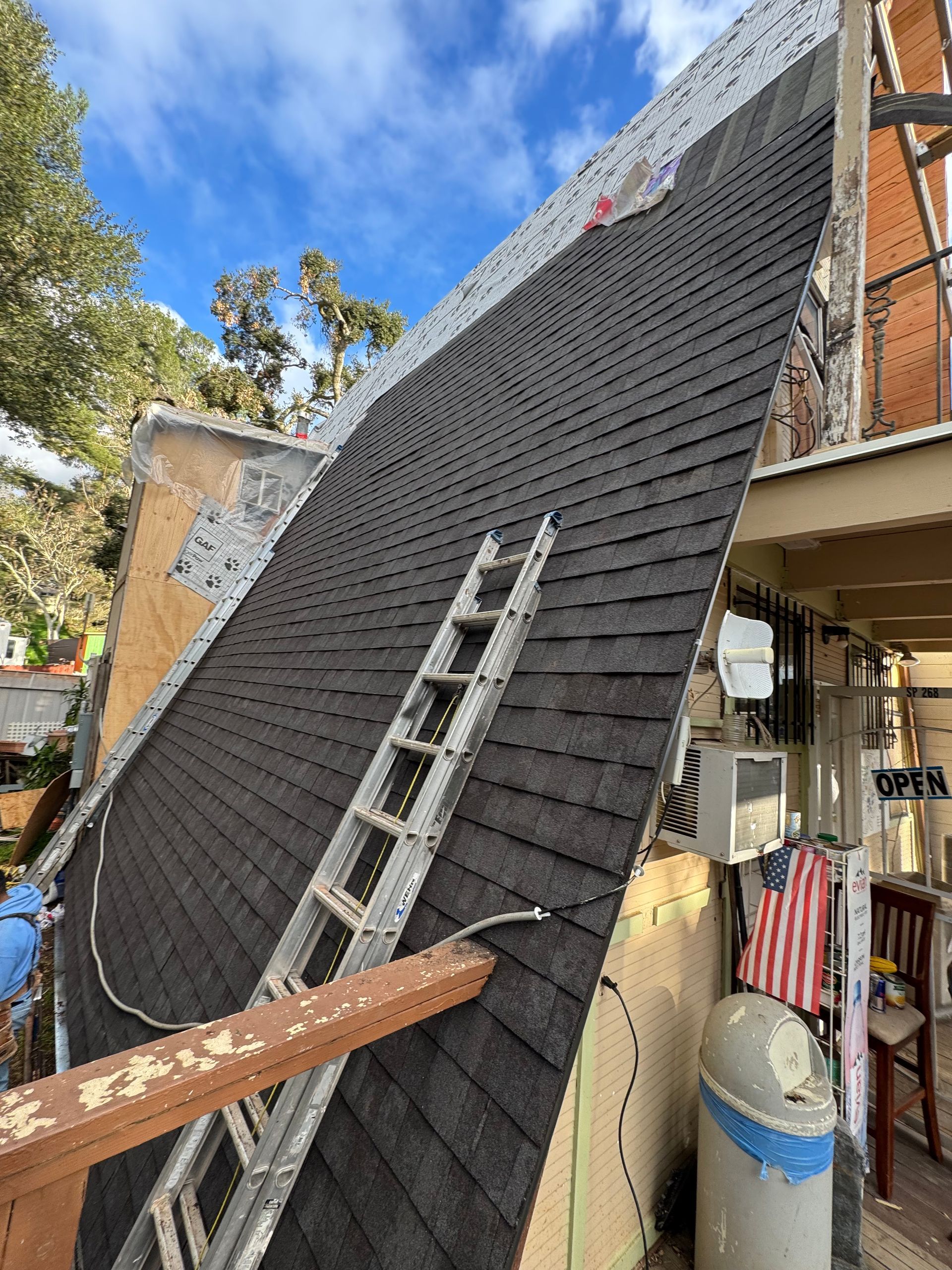 Roof partially shingled, ladder leaning against it. Dark shingles, blue sky visible.