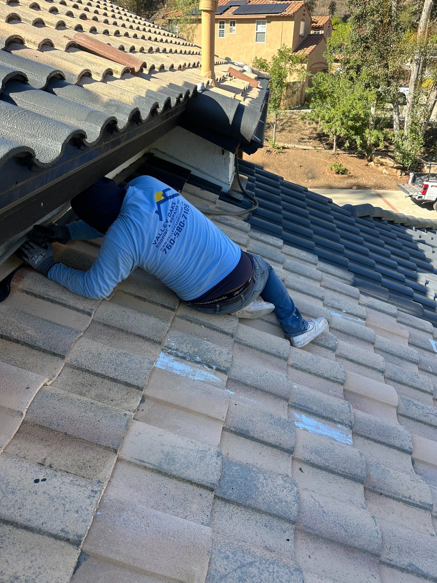 Person on a tiled roof, inspecting a gutter, wearing a blue shirt and jeans.