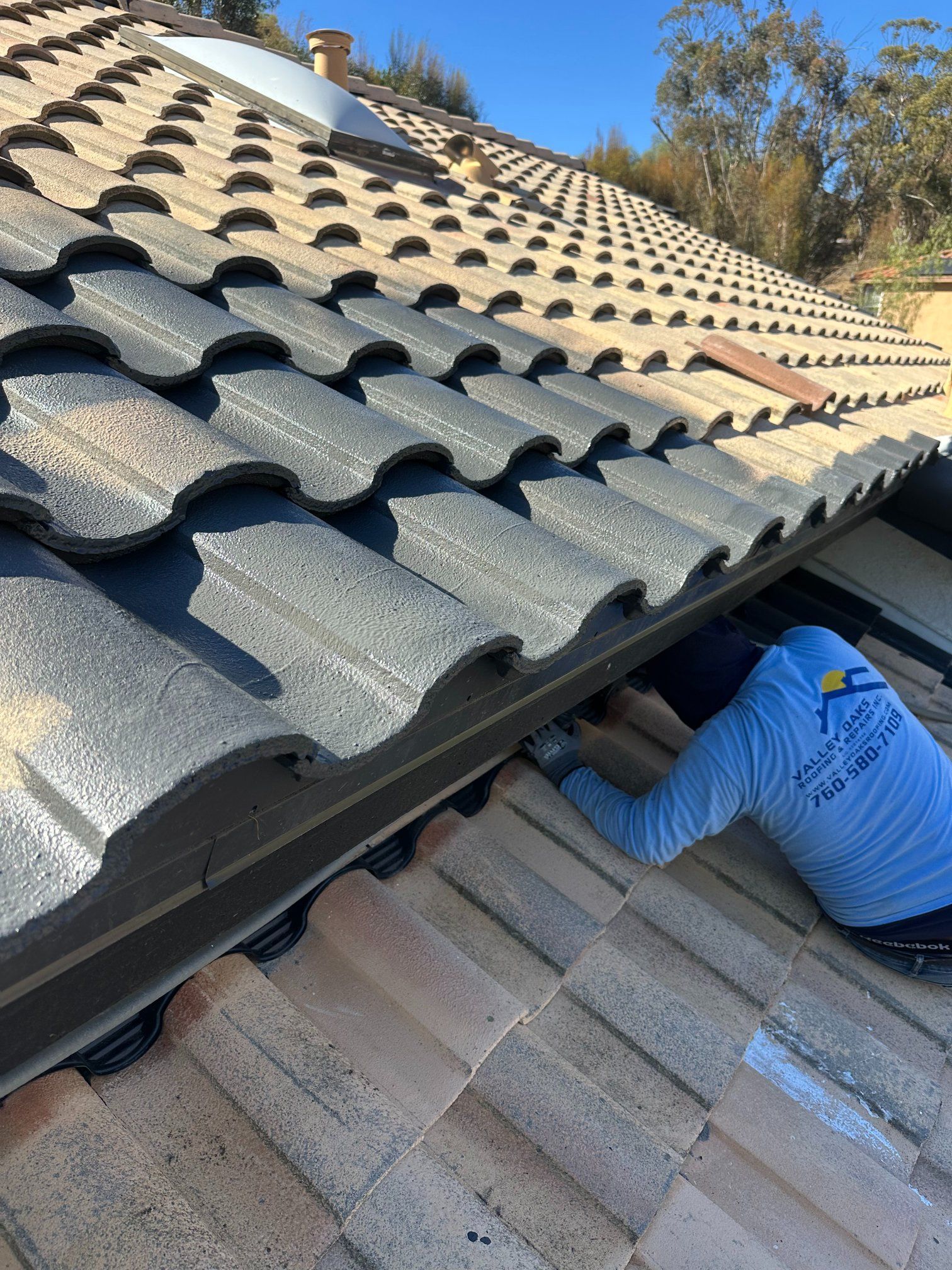 Person in blue shirt installing gutter on a roof with beige and black tiles, sunny day.