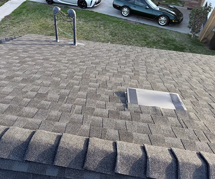Close-up of a new shingled roof with a vent and two cars parked on the lawn below.