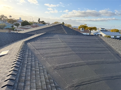 Gray roof with shingles, flat section overlaid with dark material. Houses and sky in the background.