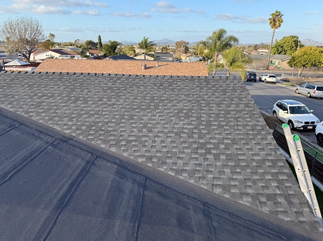 Rooftop with new gray shingles, partially completed, with a ladder, cars, and residential neighborhood.