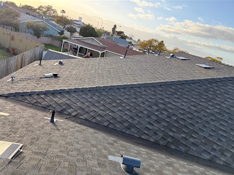 View of asphalt shingled roofs in a neighborhood. Buildings, fences, and a partly cloudy sky are in the background.