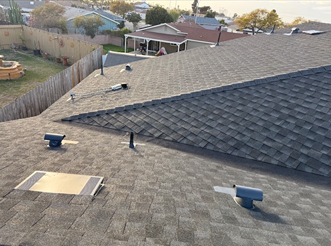 A rooftop with asphalt shingles, vents, and a view of a suburban backyard with a fence and houses.