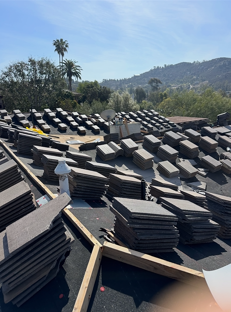 Roof under construction, stacked gray tiles on a sunny day, hills and trees in the background.