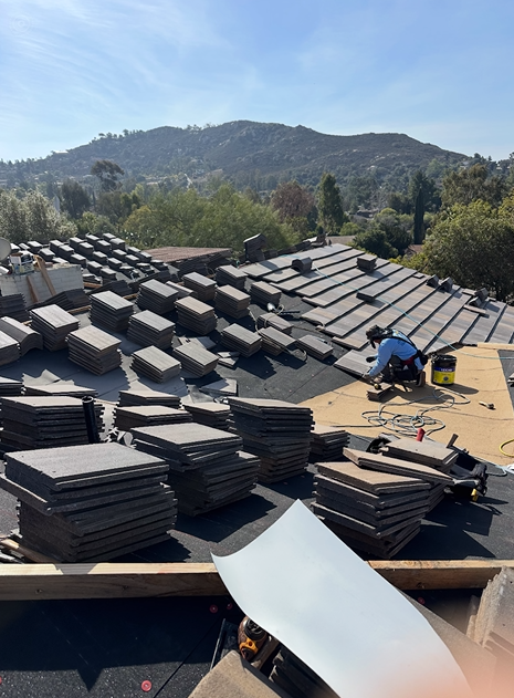 Roofer working on a tiled roof with stacks of tiles and a mountainous background.