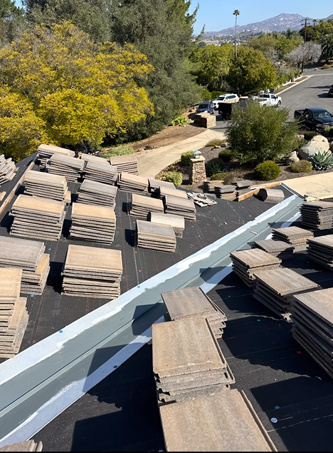 Roof tiles stacked on a roof with exposed underlayment, overlooking a driveway and trees.