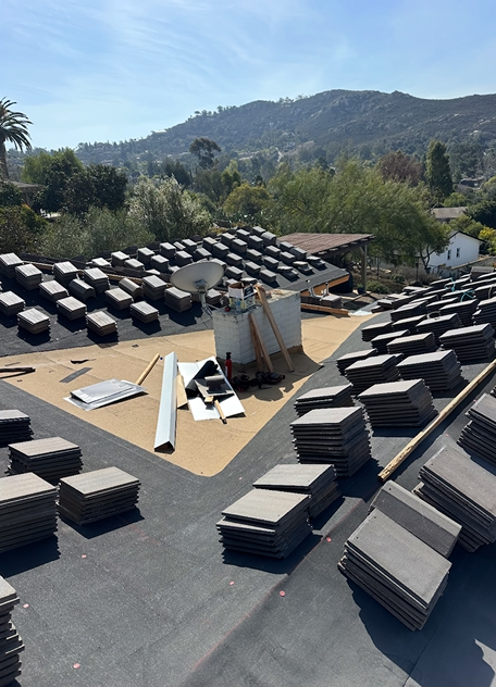 Rooftop with stacks of gray tiles being installed, chimney visible, hillside in background.