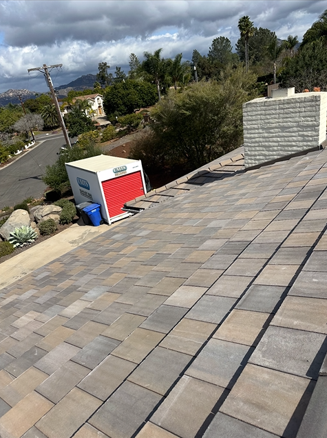 Storage container on a roof with a brick chimney and blue trash can in the sun. Road and trees in the distance.