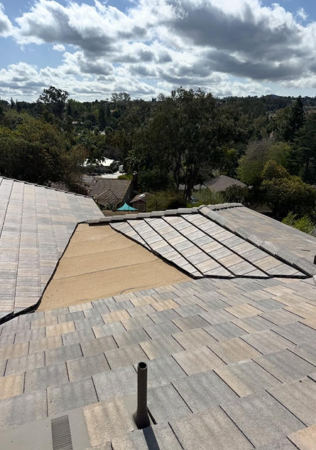 Rooftop with missing shingles and exposed underlayment; trees and houses in background under a partly cloudy sky.