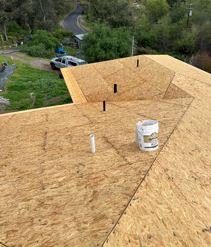 A partially constructed roof with exposed OSB sheathing, bucket, and vents. A vehicle is parked nearby in a rural setting.