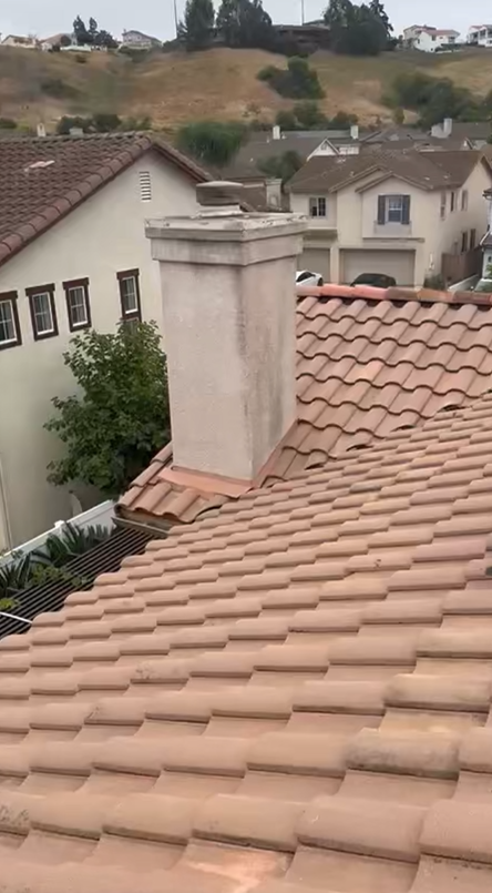 Rooftop view: red tiled roof, beige chimney, houses, green hill in background.
