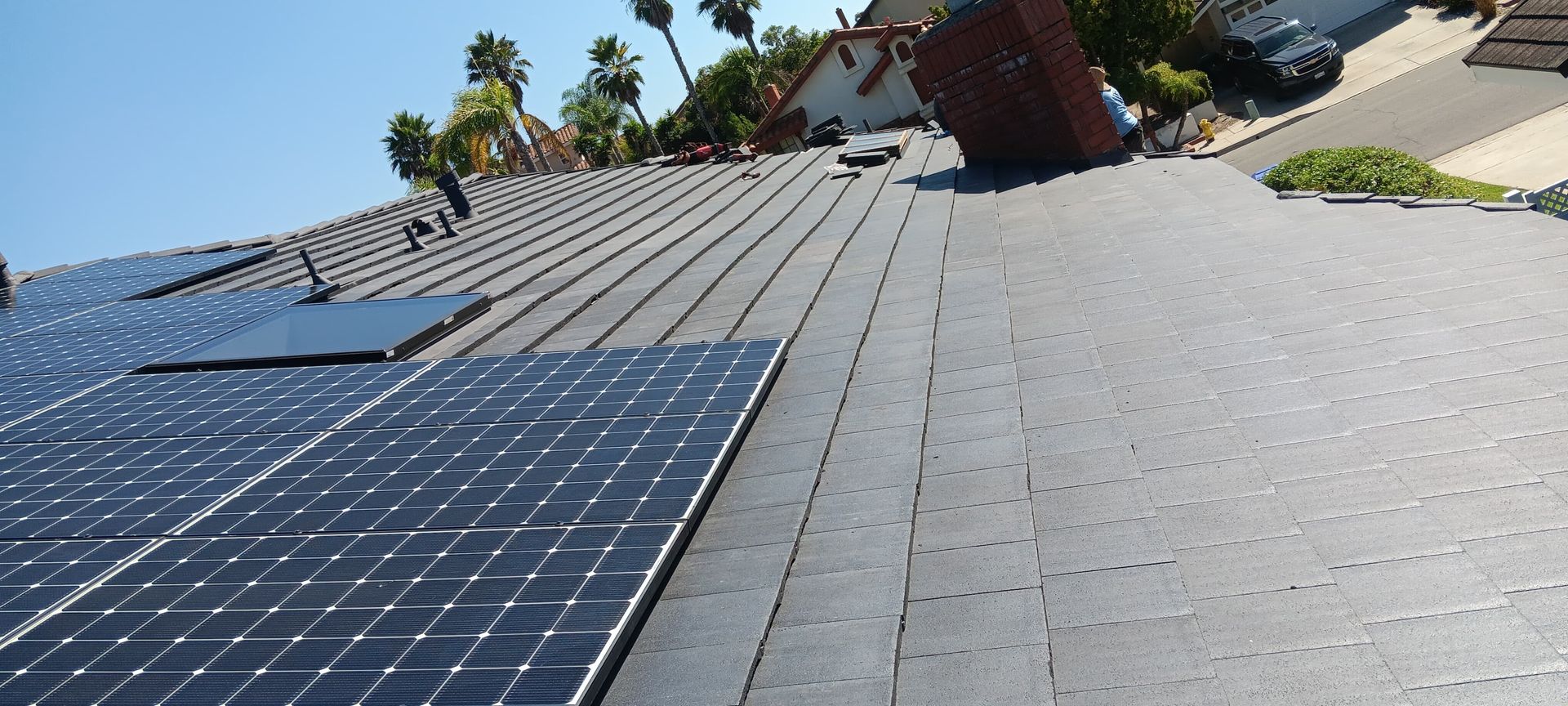 Solar panels installed on a rooftop with a chimney, blue sky, and palm trees in the background.