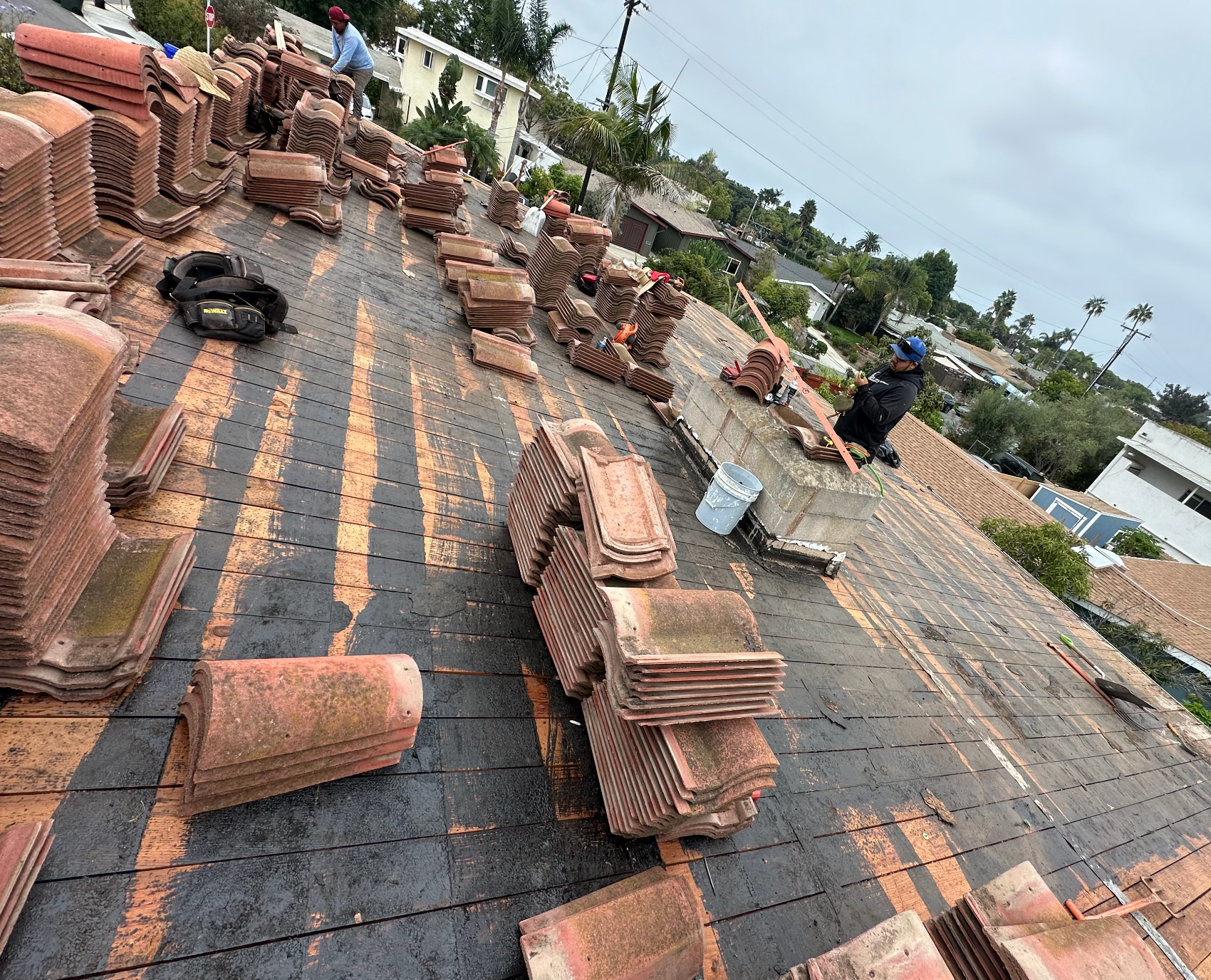 Workers replacing roof tiles on a residential building, tiles are stacked in piles; cloudy day.