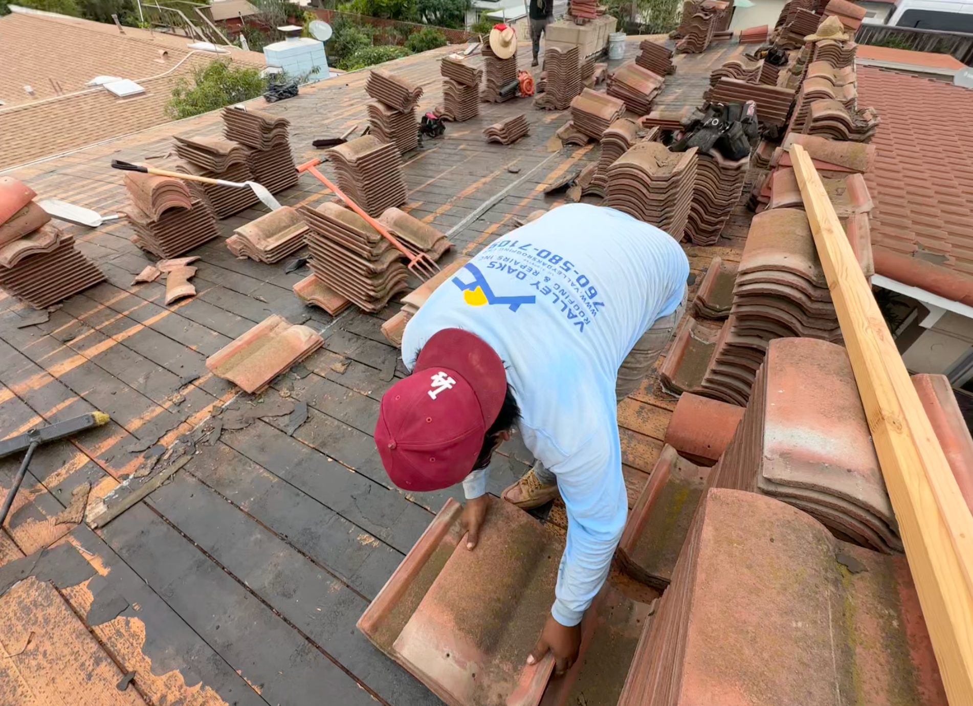 Roofer laying clay tiles on a roof. Man in blue shirt and red cap, surrounded by tiles, working outdoors.