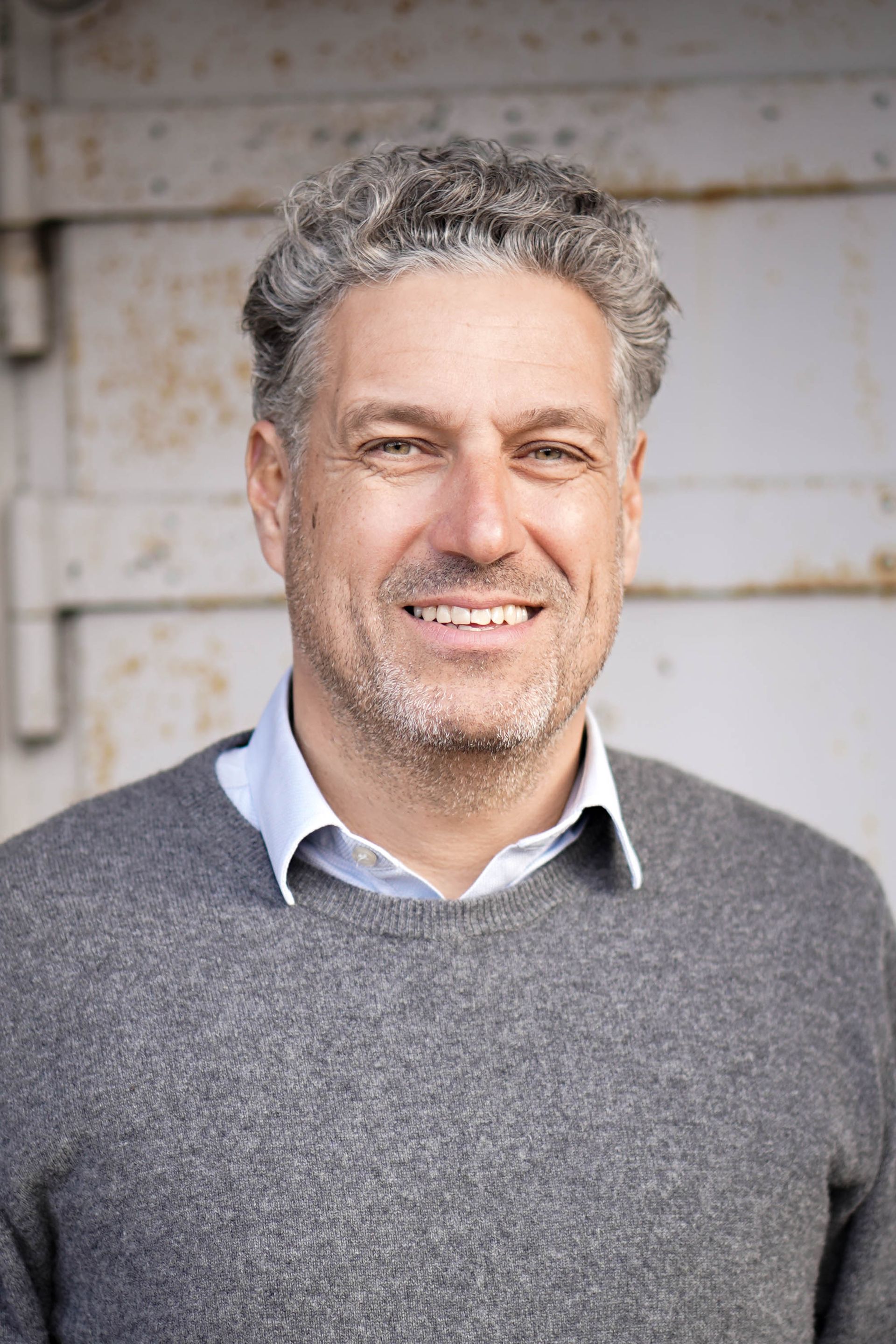 En mand med gråt hår og skæg er iført en grå sweater og smiler.