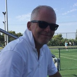 En mand iført solbriller og hvid skjorte sidder foran en tennisbane.