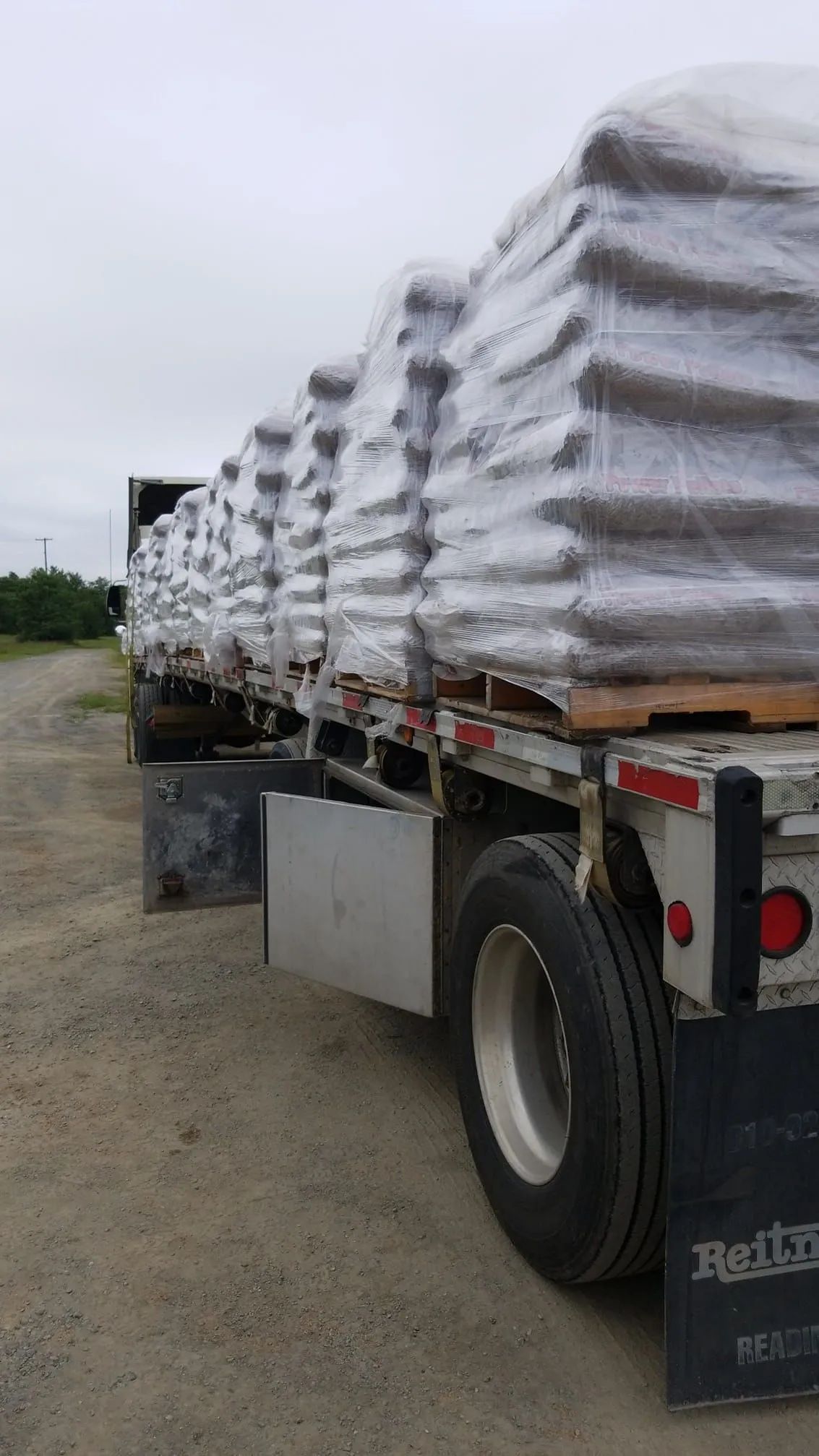 A flatbed trailer loaded with several large, shrink-wrapped pallets parked on a dirt lot under an overcast sky.