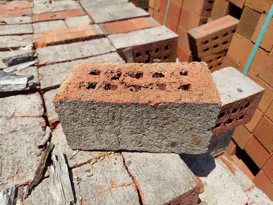 A single reddish-brown hollow brick sits on a pile of weathered, gray concrete bricks in an outdoor setting.