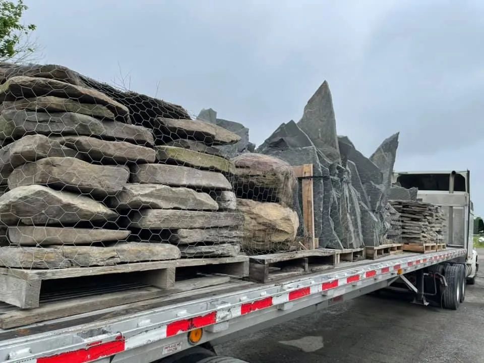 A flatbed truck loaded with stacked pallets of gray and brownish natural stone slabs and upright landscape rocks.