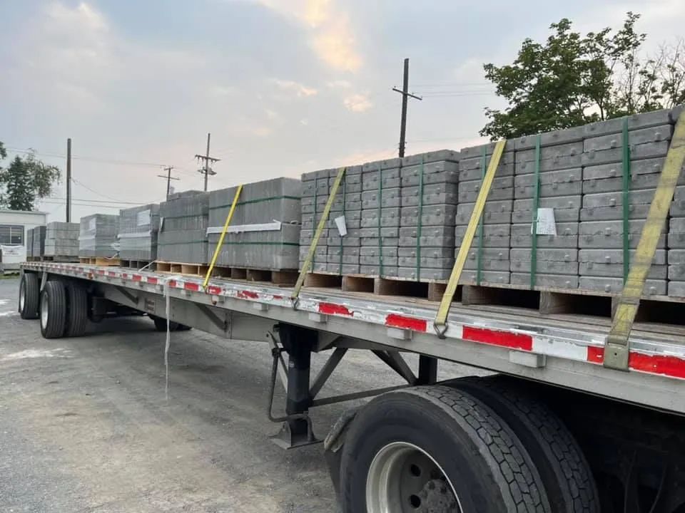 A flatbed trailer loaded with several stacks of rectangular gray concrete pavers secured by yellow ratchet straps.