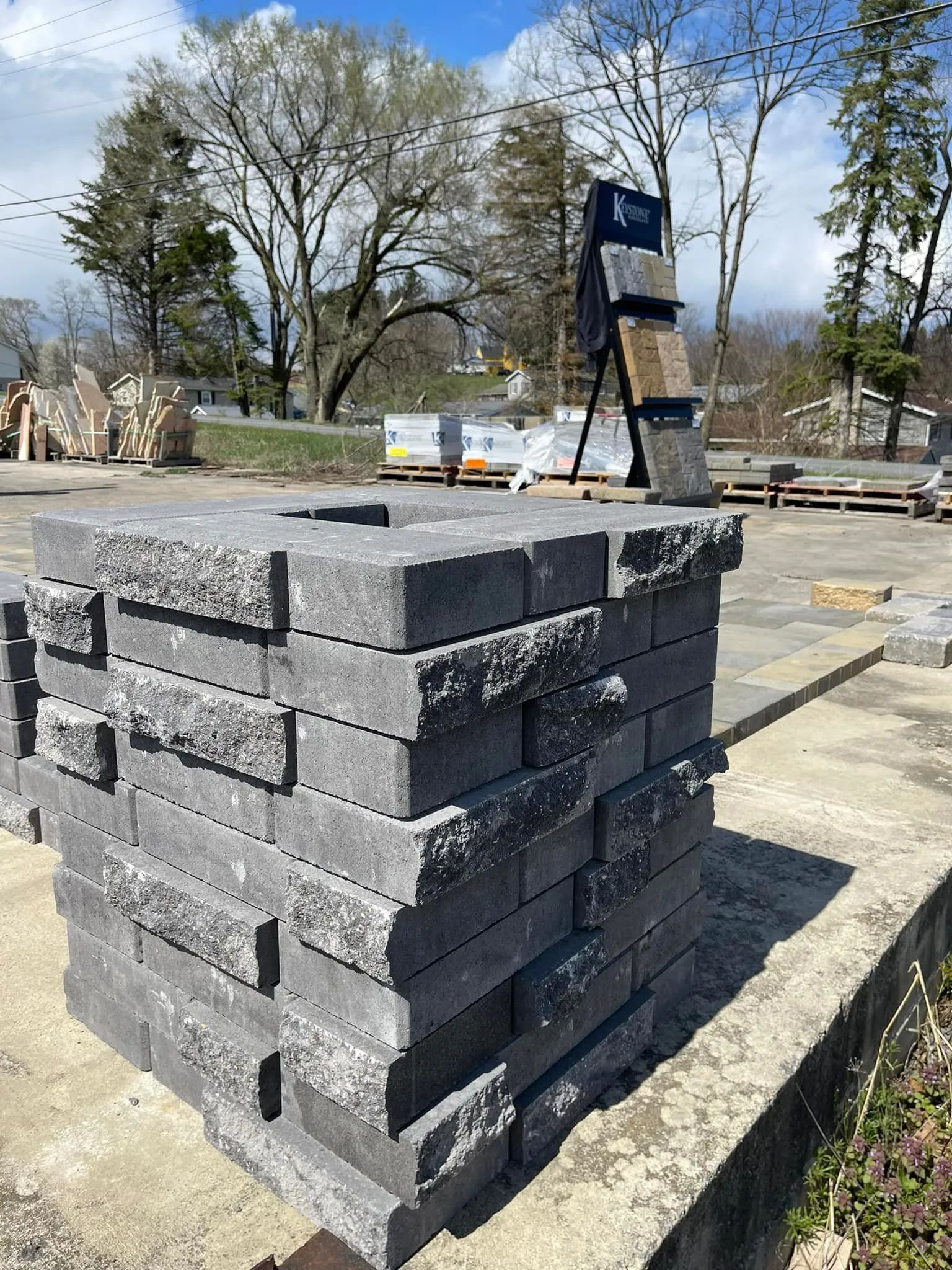 A stacked rectangular column of textured dark grey landscape blocks stands on a concrete pad outdoors.