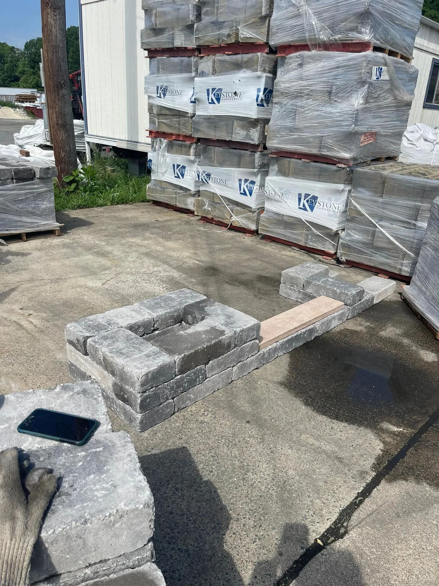 A partial masonry wall under construction near a stacked pallet of stone blocks outdoors on a gravel lot.