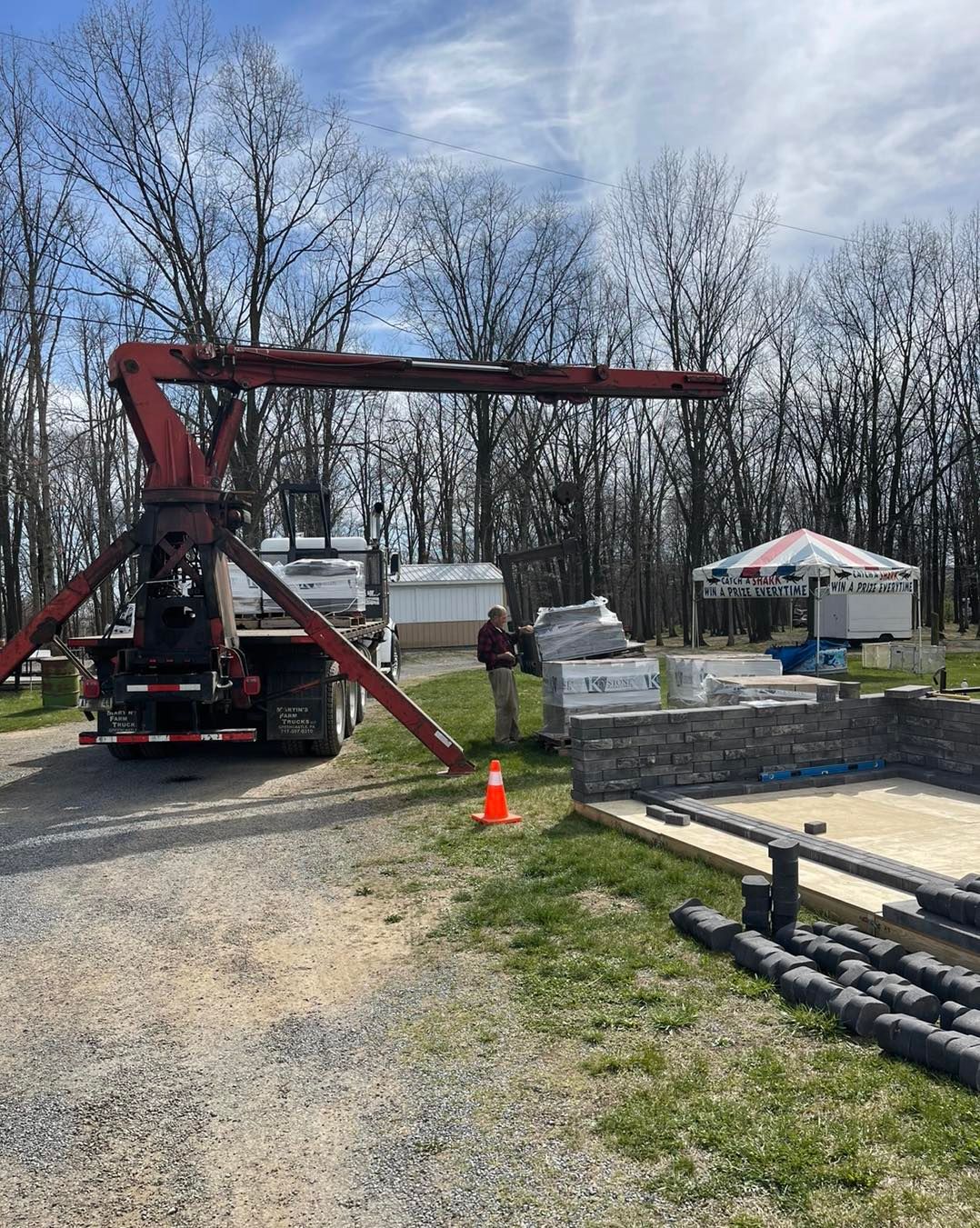 A truck with a red boom crane is parked next to stacked stone masonry supplies and a foundation on a sunny, wooded lot.