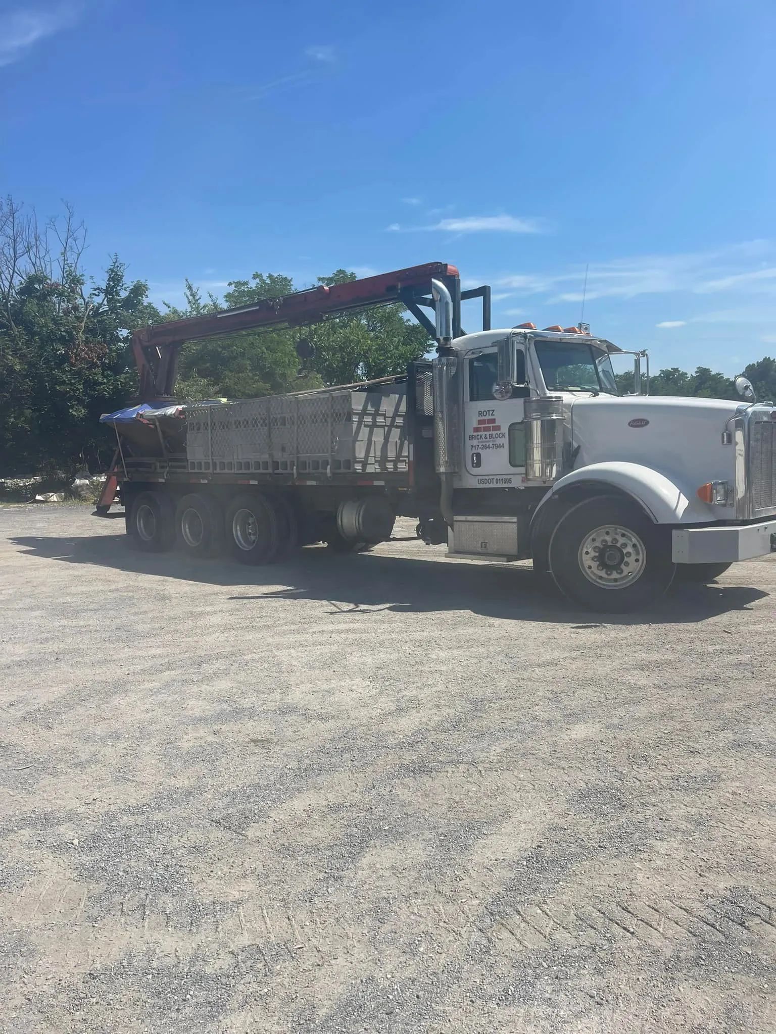 A white dump truck with a mechanical arm parked on a gravel lot under a bright blue sky.