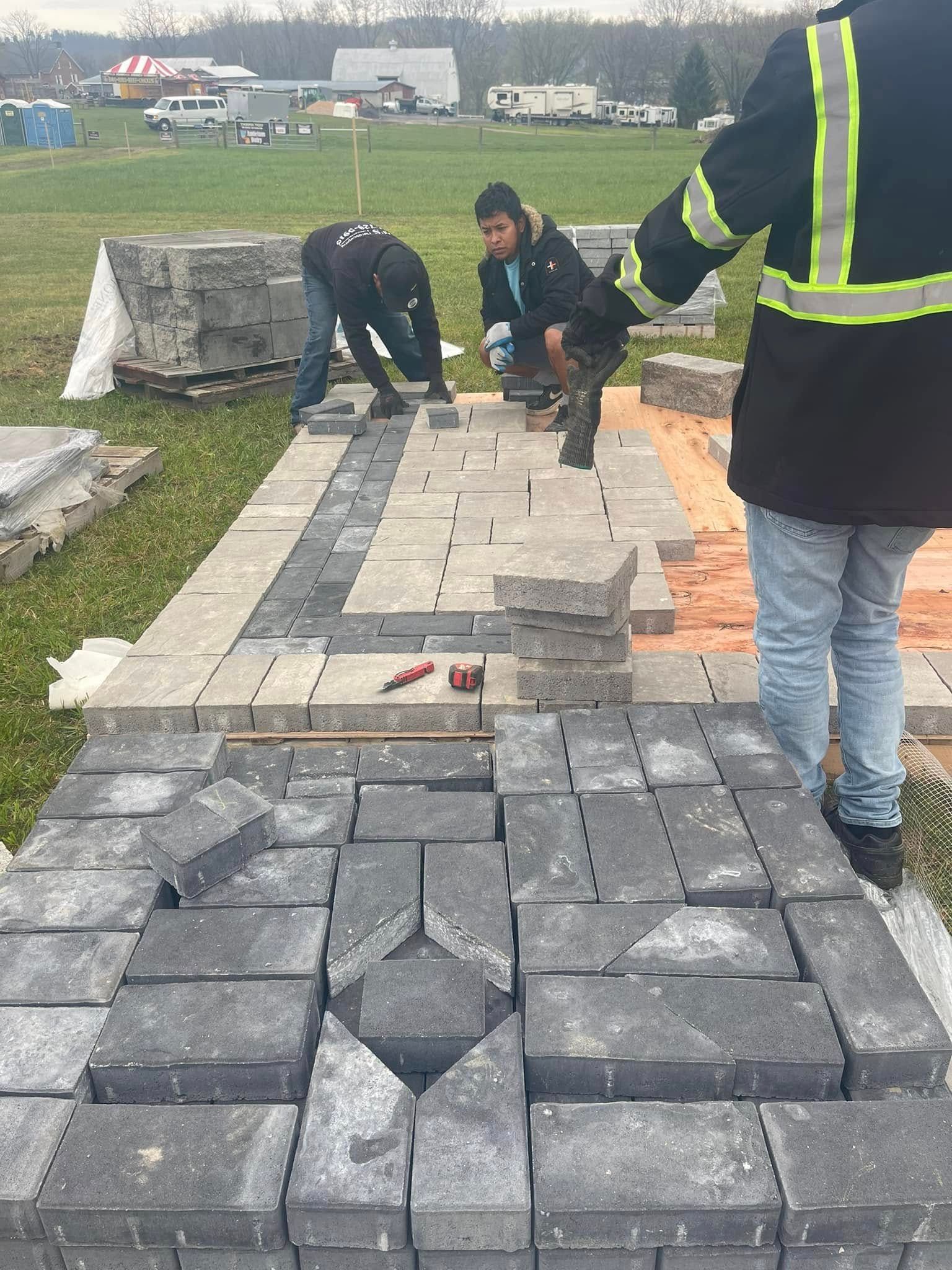 Three people installing a gray stone paver patio on a grass field, with stacks of building materials in the background.