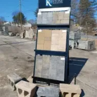 A crane lifts a pallet-moving attachment above a flatbed truck loaded with wrapped cargo, while a person stands nearby.