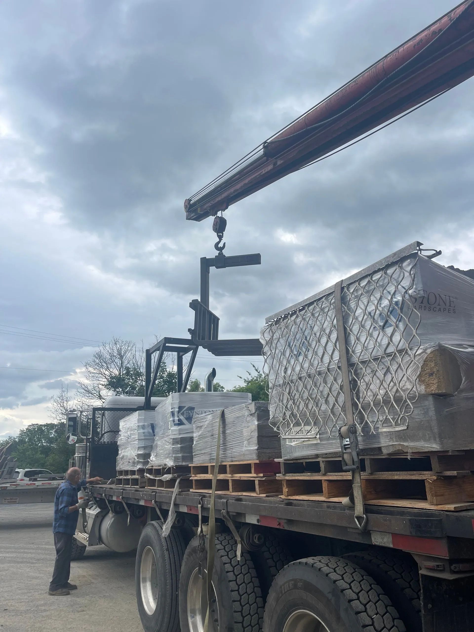 A crane lifts a pallet of stone bricks from the flatbed of a parked semi-truck under a cloudy sky.