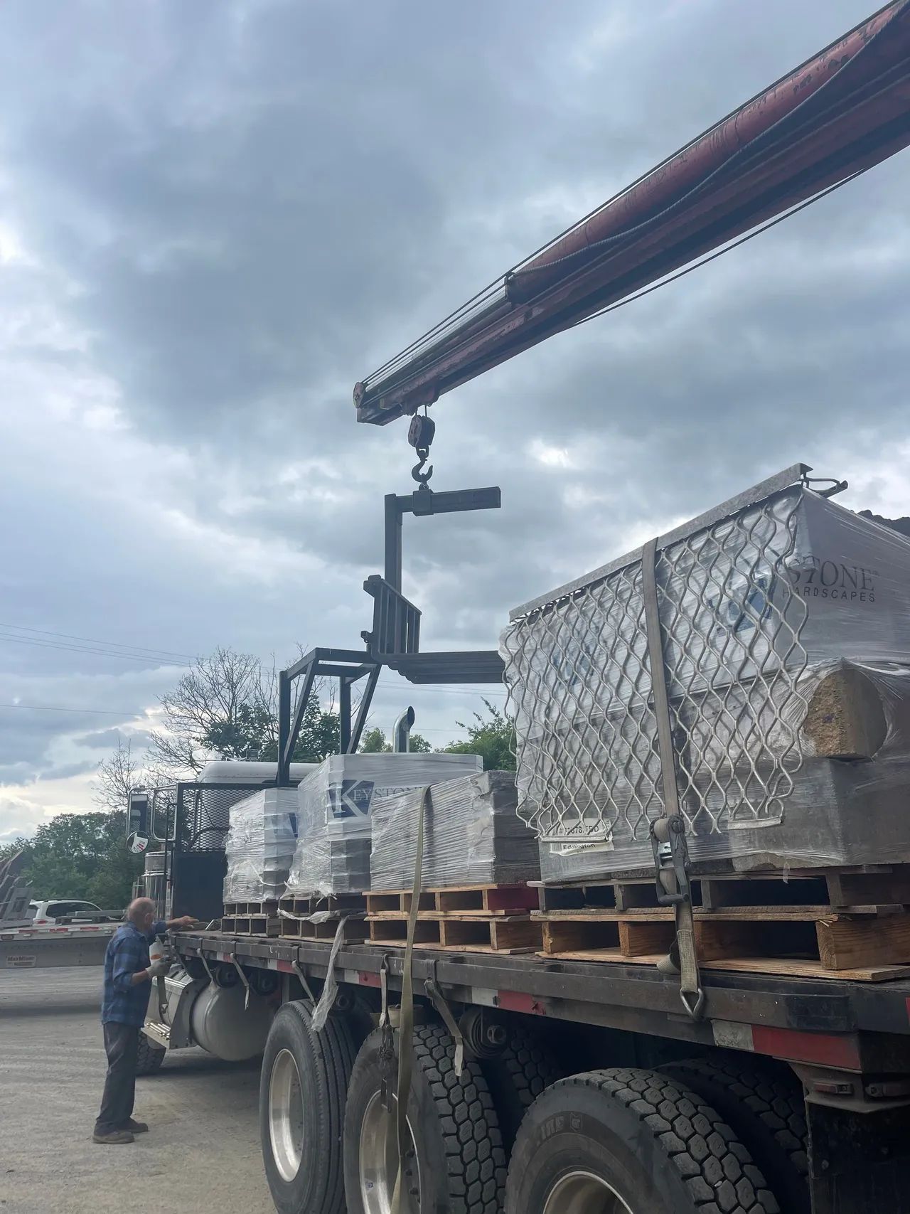 A crane attached to a truck lifts a pallet of wrapped goods while a worker stands nearby.